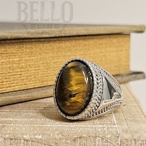 May include: A silver ring with a large oval-shaped tiger's eye gemstone. The ring has a textured band with a decorative design.