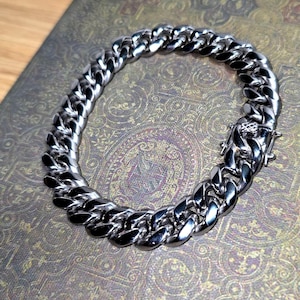 Puede incluir: Un brazalete de cadena de eslabones de color plateado. El brazalete está hecho de eslabones redondeados entrelazados y presenta un cierre seguro. La joya se exhibe sobre una superficie estampada, mostrando su acabado pulido y diseño.