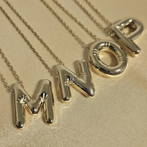 Può includere: Collane color oro con le lettere M, N, O e P. Ogni lettera è un ciondolo su una catena sottile. I ciondoli hanno un design arrotondato a bolla. Le collane sono esposte su una superficie dorata e strutturata.