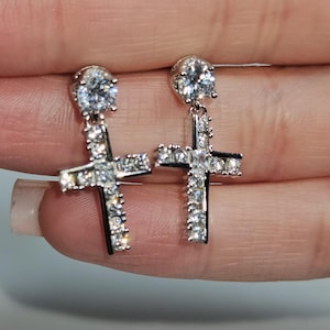 May include: A pair of silver-toned cross earrings. Each earring features a large, round, clear gemstone at the top, with a cross shape below, encrusted with smaller, clear gemstones. The crosses have a black outline.