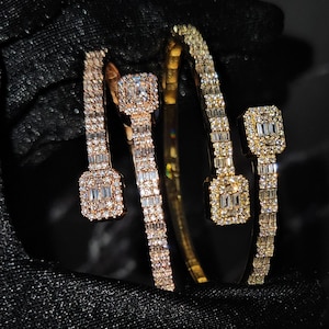 Op de afbeelding: Close-up van drie diamanten armbanden. Twee zijn roségoud, en één is goud. Elke armband heeft een reeks kleine, sprankelende diamanten en een grotere vierkante diamant in het midden. De armbanden worden op een zwart oppervlak getoond.