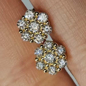 May include: A pair of gold-toned stud earrings, each featuring a cluster of clear, sparkling gemstones arranged in a floral pattern. The earrings have a small, round gold accent between each gemstone, creating a delicate and elegant design.