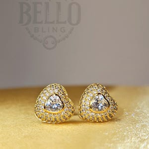 May include: A pair of gold heart-shaped earrings with a clear crystal center and smaller crystals surrounding the center. The earrings are on a gold background.