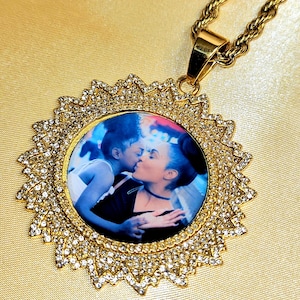 May include: Gold-toned pendant necklace featuring a circular photo of two people kissing, surrounded by a sunburst design encrusted with small, clear crystals. The chain is a thick, twisted rope style.
