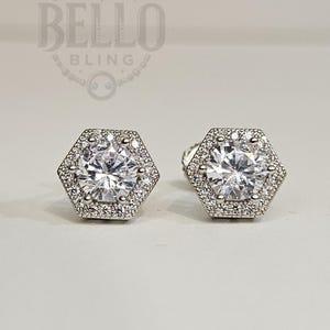 May include: A pair of silver stud earrings with a hexagonal design. Each earring features a large, clear round stone surrounded by a halo of smaller stones.