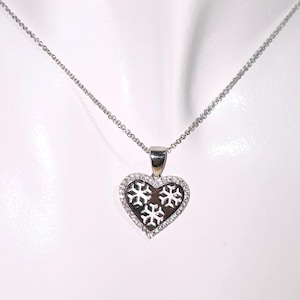 May include: A silver necklace with a heart-shaped pendant. The pendant has a dark centre with three snowflake designs and is outlined with small, clear gemstones. The necklace chain is delicate and silver.