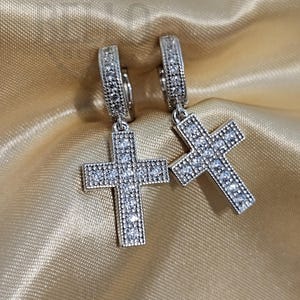 May include: A pair of silver hoop earrings with a dangling cross charm. Each cross is encrusted with clear cubic zirconia stones.