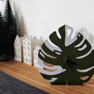 May include: A green and gray felt monstera leaf clock. The clock is standing on a wooden surface with small white houses in the background.
