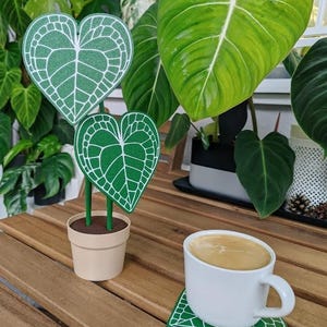 以下が含まれることがあります： 白い縁取りのある緑色のハート型の葉を持つ植物が茶色の鉢に入っています。白いマグカップにコーヒーが入っていて、緑色のハート型のコースターの上に置かれています。
