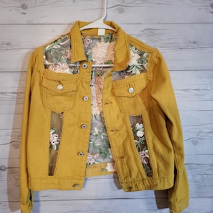 Pode incluir: Uma jaqueta de ganga amarela mostarda com uma sobreposição de renda floral. A renda é visível através do denim, criando um visual único e elegante.