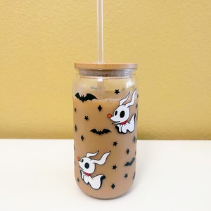 May include: A clear glass tumbler with a bamboo lid and a clear straw. The tumbler is decorated with white cartoon ghost dogs, black bats, and stars. The glass is filled with a light brown liquid.