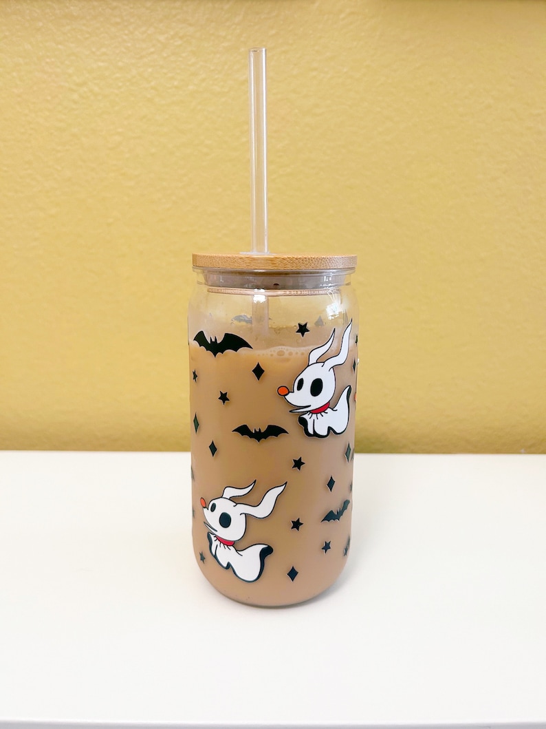 May include: A clear glass tumbler with a bamboo lid and a clear straw. The tumbler is decorated with white cartoon ghost dogs, black bats, and stars. The glass is filled with a light brown liquid.