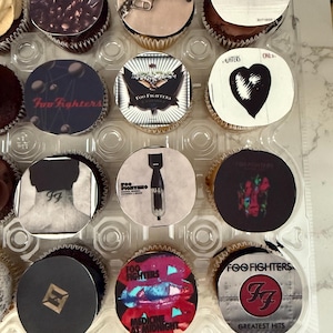 Puede incluir: Primer plano de una variedad de cupcakes decorados con diferentes portadas de álbumes de Foo Fighters. Los cupcakes están dispuestos en un recipiente de plástico transparente. Las portadas de los álbumes incluyen "Foo Fighters", "One By One", "Medicine At Midnight" y "Greatest Hits".