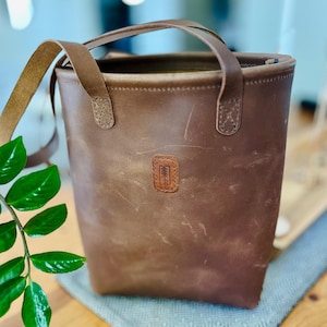 Könnte beinhalten: Eine braune Ledertasche mit einem oberen Griff und einem Schultergurt. Die Tasche hat eine rechteckige Form mit einem kleinen, rechteckigen Aufnäher auf der Vorderseite. Der Aufnäher hat ein Baumdesign. Die Tasche ist aus Leder.