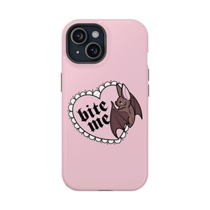 Puede incluir: Una funda de teléfono rosa con un diseño en forma de corazón y la frase "bite me" en fuente gótica. Un murciélago marrón con las alas extendidas está dentro del corazón. La funda tiene un acabado brillante y un recorte para la cámara.