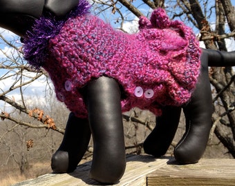Puede incluir: Un suéter de perro de punto rosa y morado con un cuello con volantes y dos botones rosas. El suéter está en un perro negro.