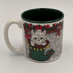 Puede incluir: Taza de cerámica blanca con interior y asa verdes. La taza presenta un diseño temático navideño con un gatito gris asomando por una media verde. El diseño incluye una chimenea, adornos y un lazo rojo.