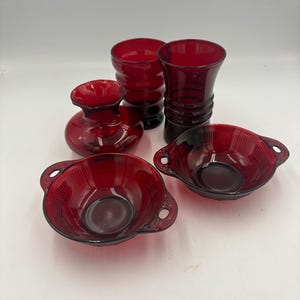 May include: A collection of five ruby red glass items including two small bowls with side handles, a rounded vase with a ruffled rim, a ribbed cylindrical vase, and a tall ribbed tumbler. Each piece features a deep, translucent red color.