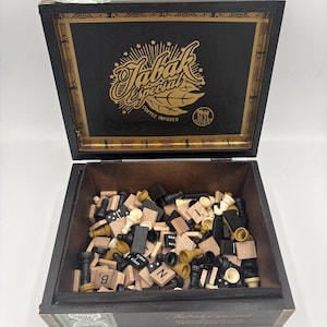 Puede incluir: Una caja de madera negra con tapa con bisagras, llena de piezas de juego variadas. La tapa tiene letras doradas que dicen "Tabak Especial Coffee Infused". En el interior hay piezas de ajedrez, dominó y fichas de letras. La caja está abierta.
