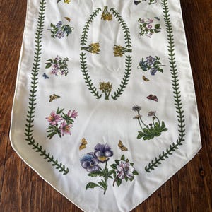 Puede incluir: Camino de mesa floral blanco con un diseño botánico. El camino de mesa presenta varias ilustraciones de flores coloridas, mariposas y un borde de hojas verdes. Las flores incluyen flores rosas, moradas y amarillas.