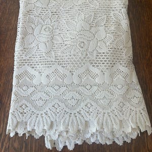 May include: Cream-colored lace tablecloth with intricate floral and geometric patterns. The tablecloth is folded, showcasing the detailed openwork design and scalloped edge. The fabric appears to be cotton or a similar material, suitable for a dining table.