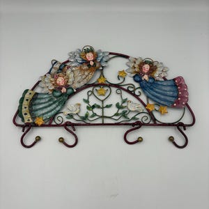 Op de afbeelding: Een decoratieve metalen wandhaak met drie engelenfiguren met bloemen vleugels en halo's. De engelen zijn geschilderd in pastelkleuren en zijn gerangschikt boven drie gebogen haken. Het stuk is ongeveer 30 cm breed.