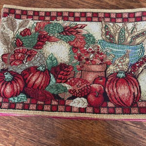 Puede incluir: Un mantel individual rectangular de tapiz tejido con un diseño de cosecha que presenta calabazas rojas, hojas verdes, una maceta de terracota y un cuenco azul lleno de maíz. El borde tiene un patrón de cuadros rojos y beige.