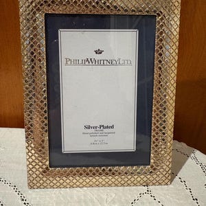 May include: A silver-plated picture frame with a textured, gold-coloured border. The frame holds a dark blue matting and a white insert with the text "PHILIPWHITNEY LTD" and "Silver-Plated FRAME" with dimensions of 8.8cm x 12.7cm.