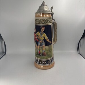 May include: A tall ceramic beer stein with a pewter lid and handle. The body features a relief illustration of two soccer players on a field, with the text "SPIEL-TRINK-NIE-ZU" printed in black lettering along the base of the decorative scene.