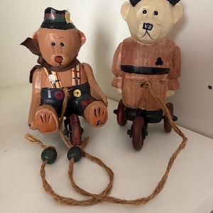 Puede incluir: Dos figuras de osos de madera sobre pequeños vehículos con ruedas. Uno de los osos lleva sombrero y tirantes, el otro un abrigo. Ambos tienen correas de cuerda marrón con cuentas verdes. Los osos están pintados en tonos marrones, negros y azules.