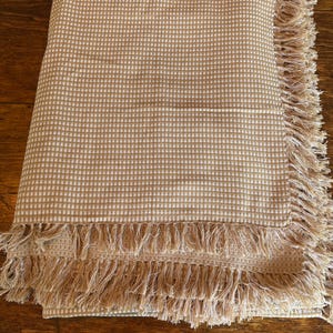 May include: A folded rectangular throw blanket featuring a small-scale tan and white chequered pattern. The fabric has a textured weave and is finished with long, loose fringe detailing along two of the edges, creating a soft, layered appearance.