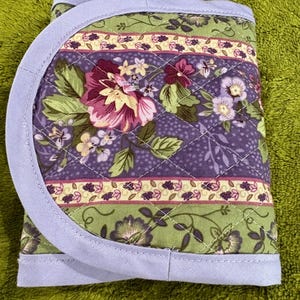 May include: A quilted fabric item with a floral pattern in shades of purple, green, and pink. The item has a light blue border and a diamond-stitched design. The floral pattern includes various flowers and leaves.