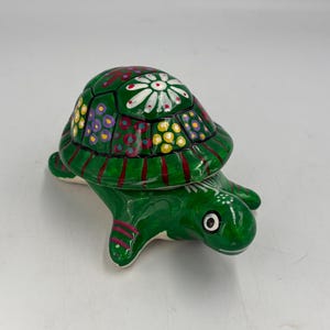 Könnte beinhalten: Eine grüne Keramik-Schildkrötenfigur mit handbemaltem Panzer, der mit weißen, gelben und violetten Blumenmustern verziert ist. Die Schildkröte hat eine glänzende Oberfläche, ein weißes Auge mit schwarzer Pupille und rote Streifen an den Flossen.