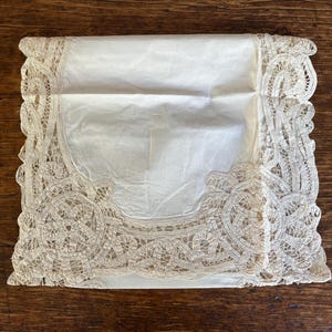 May include: A folded white linen handkerchief with intricate, cream-colored lace trim. The lace forms a decorative border around the edges, adding a vintage and elegant touch. The handkerchief is placed on a dark wooden surface.
