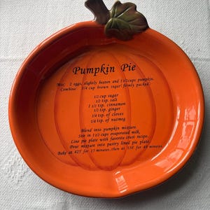 May include: A bright orange ceramic pie dish shaped like a pumpkin with a green stem and leaf handle. The interior features the text 'Pumpkin Pie' followed by a recipe listing ingredients like eggs, pumpkin, sugar, salt, cinnamon, ginger, cloves, nutmeg, and evaporated milk, along with baking instructions.