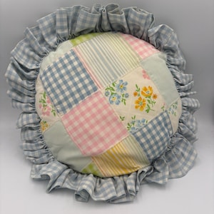 May include: A round patchwork pillow featuring a variety of fabric squares in pastel colors including light blue, pink, yellow, and green. The patterns include gingham checks, stripes, and floral prints. A ruffled light blue gingham border surrounds it.
