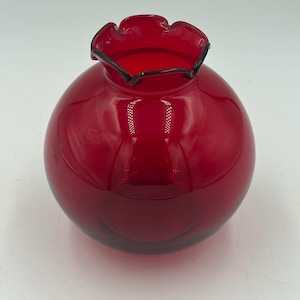 May include: A small, spherical ruby red glass vase with a smooth, glossy finish. The opening at the top features a distinct ruffled, crimped edge design, creating a decorative, wavy rim that contrasts with the rounded, bulbous body of the vessel.