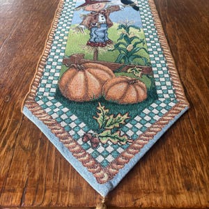 May include: A rectangular woven table runner featuring a scarecrow, pumpkins, corn stalks, and a black bird on a light blue background. The border has a green and white checkered pattern with a tan rope trim and a gold tassel at the pointed end.
