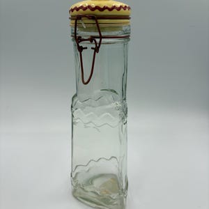 May include: A clear glass jar with a decorative yellow and red lid. The jar has a rectangular shape with a wavy design on the sides and a metal clasp closure. The lid has a red knob.