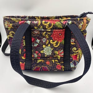 Puede incluir: Bolso tote acolchado con un estampado floral en rojo, amarillo y azul sobre un fondo oscuro. El bolso tiene asas azul marino y cierre de cremallera. El bolso mide aproximadamente 30 cm de ancho y 23 cm de alto.