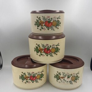 May include: A set of four round, cream-colored storage containers with dark brown lids. Each container features a printed graphic of a red tomato, a yellow onion, green peppers, and mushrooms surrounded by green foliage and small red berries.