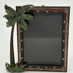May include: Decorative photo frame with a tropical theme. The frame is rectangular with a brown and bronze finish, accented with small rhinestones. A detailed palm tree and grass adorn the corner, adding a touch of nature to the design.