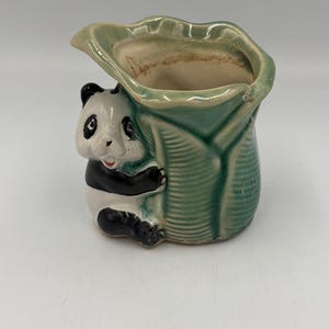 Puede incluir: Una maceta de cerámica con forma de hoja verde y un oso panda esculpido que se aferra al lateral. El panda tiene marcas blancas y negras, y la hoja presenta un acabado brillante con nervaduras marcadas. El interior es de color beige.