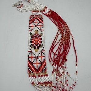 May include: A beaded necklace featuring a rectangular pendant with a turtle motif in red, white, black, and gold seed beads. The pendant is attached to multiple long strands of red, white, and gold beads, finished with a silver-toned metal clasp.