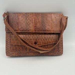 May include: A rectangular brown shoulder bag featuring a textured snakeskin pattern. The bag has a fold-over flap design, a front pocket with a single snap button closure, and a thin matching shoulder strap attached with silver-toned metal rings.