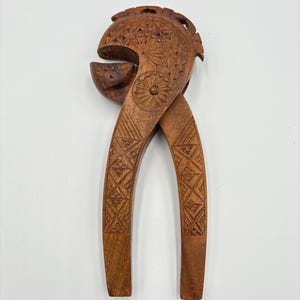 Op de afbeelding: Een vintage houten notenkraker met ingewikkelde gesneden details. De bruine notenkraker heeft een gedetailleerd bloemen- en geometrisch patroon. De handvatten zijn gekruist en de kop heeft een afgeronde vorm met een nootkrakeropening.