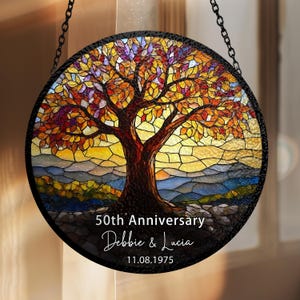 Puede incluir: Vidriera redonda con un árbol de hojas otoñales coloridas. El árbol se alza sobre un fondo de puesta de sol con tonos amarillos y azules. Texto: "50th Anniversary", nombres y una fecha.