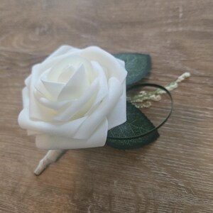 May include: A white rose boutonniere with green leaves and a stem. The flower is in full bloom, with multiple layers of petals. The leaves are a dark green color, and the stem is wrapped in a light-colored material. This floral accessory is suitable for weddings or special events.