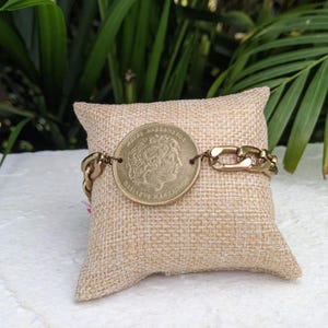 Könnte beinhalten: Ein goldfarbenes Armband mit einem großen Münzenanhänger, der das Profil einer Frau zeigt. Die Münze trägt die Aufschrift "REPUBLICA ARGENTINA" und "BALBOA MARIANO" am Rand. Das Armband liegt auf einer beigefarbenen Stoffoberfläche.