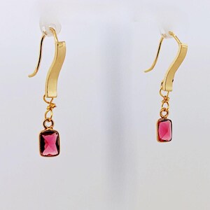 May include: A pair of gold earrings with a dangling pink gemstone. The earrings have a simple, curved design.